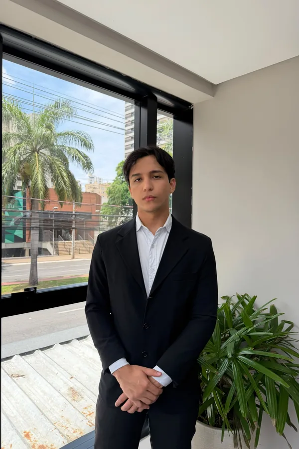 Henrryk Ribeiro Silva, assistente jurídico, de corpo inteiro, de terno preto e camisa branca, em frente a uma janela