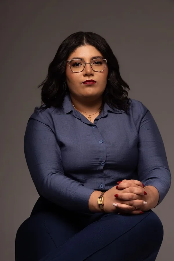 Jackeline Jesus Telles, assistente jurídica, de corpo inteiro, sentada, de óculos e camisa azul, com as mãos entrelaçadas