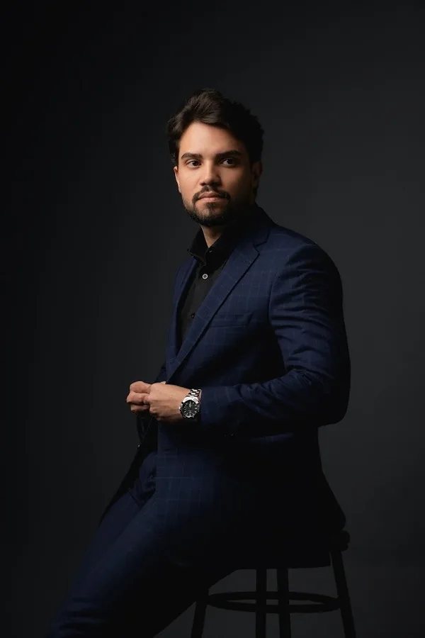 Lucas Almeida Duarte Primo, advogado, de corpo inteiro, sentado de terno azul-marinho xadrez e camisa preta, ajeitando o paletó