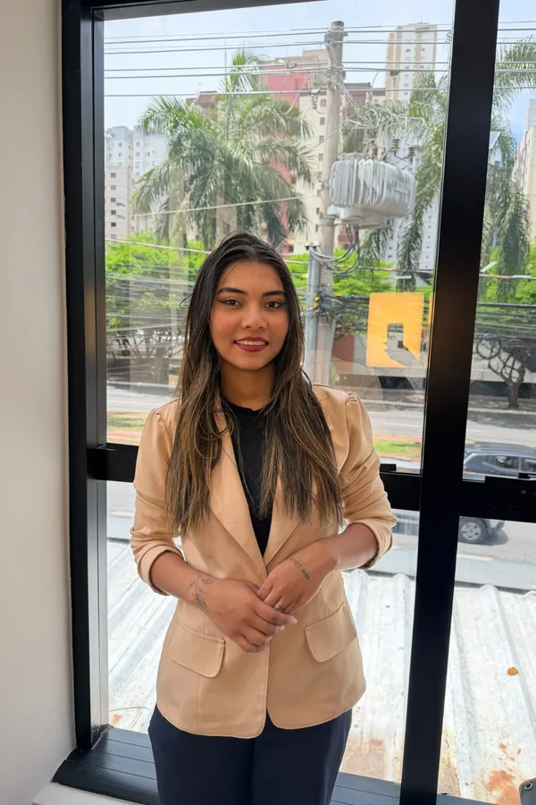 Pamela Raissa Nascimento Pinheiro, assistente jurídica, de corpo inteiro, de blazer bege, sorrindo, em frente a uma janela